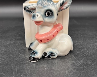 May include: A vintage ceramic donkey planter. The donkey is light blue and white with black accents, a pink scarf, and large ears. The donkey is in front of a white rectangular planter. The planter is on a dark surface.