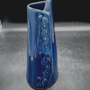May include: A tall, blue ceramic vase with a unique, asymmetrical shape. The vase features a series of embossed floral designs cascading down one side. The glossy finish and dark blue color give it a sophisticated look, suitable for home decor.