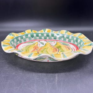 May include: Oval ceramic serving dish with a scalloped edge. The rim features a yellow and green checkered pattern, with a red stripe. The center of the dish is decorated with a painted pear design. The dish is white with green and red accents.