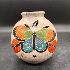 May include: A round, white ceramic vase with a butterfly design. The butterfly has green, blue, orange, and black wings. The vase has a speckled glaze and small holes throughout.