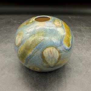 May include: A spherical ceramic vase with a glossy finish, featuring a mottled glaze in shades of blue, olive green, and tan. The round vessel has a small, circular opening at the top and a smooth, curved body with blended color transitions.