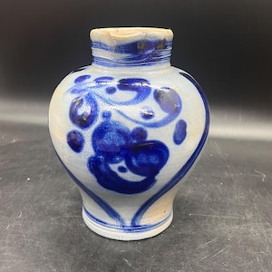 May include: A ceramic vase with a bulbous body and a narrow neck. The vase is primarily off-white with blue floral designs and accents. The rim and base feature blue bands. The vase has a rustic, handcrafted appearance.