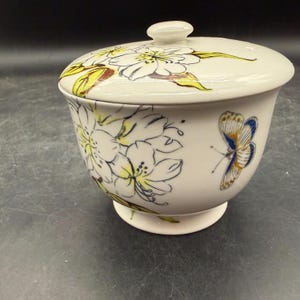May include: A white ceramic bowl with a lid, adorned with floral and butterfly designs. The bowl features a pedestal base and a rounded shape. The lid has a small knob for easy handling. The floral design includes white flowers with yellow and brown accents.