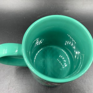 May include: A green ceramic mug with a smooth, glossy finish. The mug has a rounded handle and a wide, open mouth.