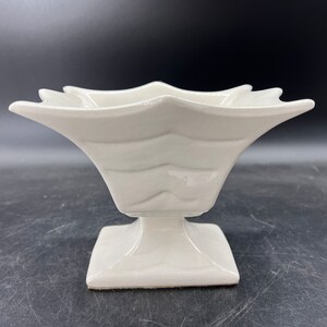 May include: A white ceramic pedestal bowl with a star-shaped rim and a fluted design. The bowl is on a square base.