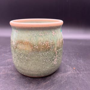 May include: A small, round, green ceramic pot with a speckled glaze. The pot has a light brown rim.