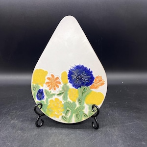 May include: A white, teardrop-shaped ceramic decorative plate with a colorful floral design. The flowers are yellow, orange, and blue, with green leaves. The plate is displayed on a black metal stand.