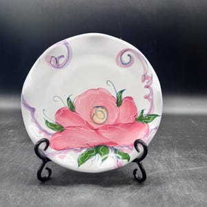 May include: A white ceramic plate with a pink flower painted on it. The flower has green leaves and a yellow center. The plate has a purple and blue swirl design around the edge.