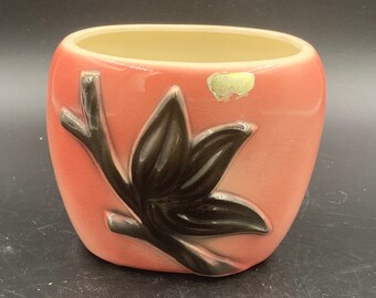 May include: A pink ceramic planter with a black stylized flower design. The planter has a slightly rounded shape and a smooth, glossy finish.