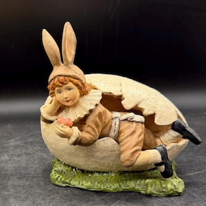 May include: A figurine of a child dressed in a bunny costume emerging from a cracked egg. The child wears a brown outfit, a bunny ear hat, and black shoes. The egg is beige and sits on a green, grassy base. The child holds a small pink flower.