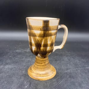 May include: A ceramic coffee mug with a pedestal base. The mug features a cream-colored interior and a brown plaid pattern on the exterior. The handle is a matching cream color. The base is a light brown color.