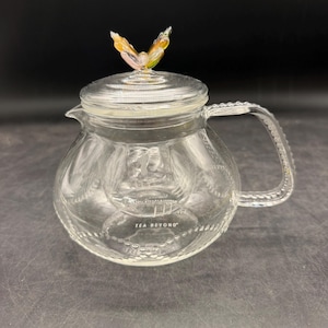 May include: A clear glass teapot featuring a rounded body with a beaded texture along the base and handle. The lid is topped with a small, multicolored glass butterfly finial. The text TEA BEYOND is printed in white on the front of the glass body.