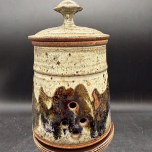 May include: A handcrafted ceramic jar with a lid. The jar is cylindrical with a textured, speckled off-white and brown glaze. It features a series of small holes near the base and a decorative finial on the lid. The jar is approximately 8 inches tall.