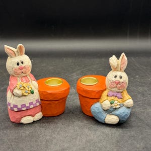 May include: Two carved wooden Easter bunny candle holders. One bunny wears a pink dress and apron, holding flowers. The other bunny wears a yellow shirt and blue pants. The candle holders are orange.