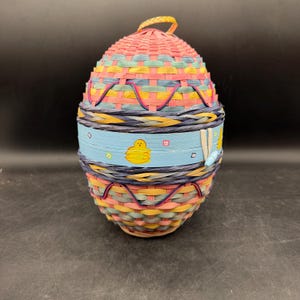May include: A woven Easter egg basket in a colorful design. The egg-shaped basket features a pink top, a blue band with a yellow chick, and a woven handle. The basket is made of woven materials in shades of pink, yellow, blue, and green.