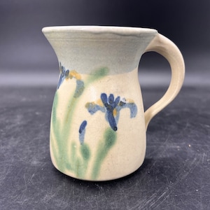 May include: A ceramic mug with a light beige glaze and a blue and green floral design. The mug has a rounded body and a handle.