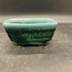 May include: A small, rectangular, green ceramic planter with a glazed finish. The planter has a slightly rounded shape and a subtle design on the front.