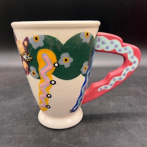 May include: A ceramic mug with a white base and a decorative handle. The mug features a green heart-shaped design with floral accents, a pink handle with blue polka dots, and a yellow and pink zigzag pattern. The mug is approximately 4 inches tall.
