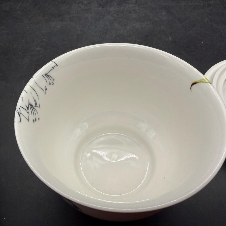 May include: A white ceramic bowl with a floral design in black ink on the rim. The bowl has a smooth, glossy finish and a rounded shape. The interior is a soft white color, and the exterior rim has a thin pink band.