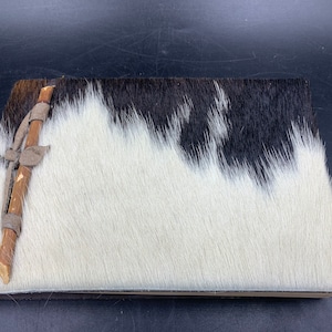 May include: A black and white cowhide journal with a wooden stick wrapped in leather running diagonally across the cover.