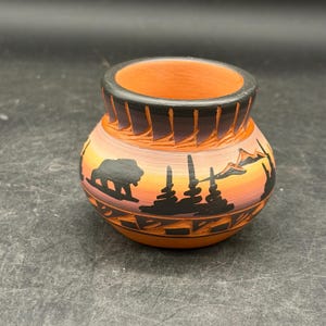May include: A small, round, terracotta-colored ceramic pot with a black rim and a landscape design. The design features a bear, trees, and mountains silhouetted against a sunset gradient of orange, pink, and yellow.