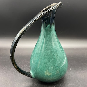 May include: A green ceramic jug with a black handle and a distinctive, curved spout. The jug has a textured, speckled glaze.