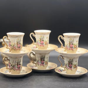 May include: Set of six floral patterned teacups and saucers with gold trim. The cups and saucers are white with a cream-coloured background and feature a delicate floral design in shades of pink, blue, and yellow.