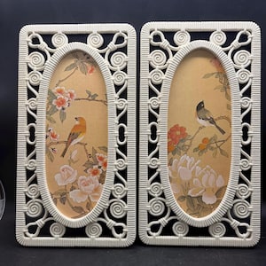 May include: Two rectangular wall plaques featuring off-white, intricate, scroll-patterned frames surrounding oval inserts. Each insert displays a light tan background with illustrations of birds perched on flowering branches with pink and white blossoms.