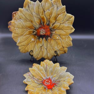 May include: Two ceramic dishes shaped like flowers with brown and yellow petals and a red center. The larger dish is on a stand.