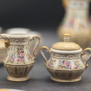 May include: A vintage porcelain creamer and sugar bowl set with floral designs and gold accents. The creamer has a handle and a spout, while the sugar bowl has a lid and two handles.