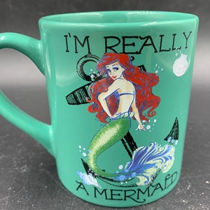 May include: A green ceramic mug with a white handle. The mug features a graphic of Ariel, the Little Mermaid, with an anchor and the text "I'm Really A Mermaid".