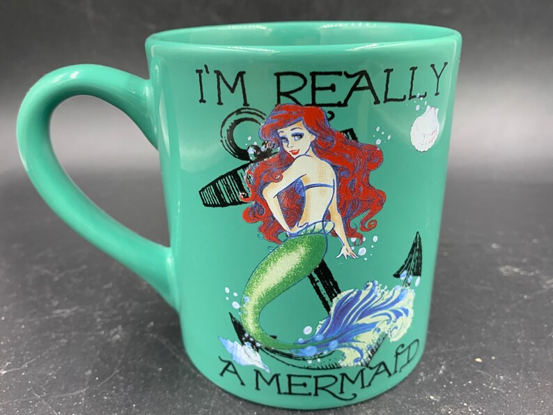 May include: A green ceramic mug with a white handle. The mug features a graphic of Ariel, the Little Mermaid, with an anchor and the text "I'm Really A Mermaid".
