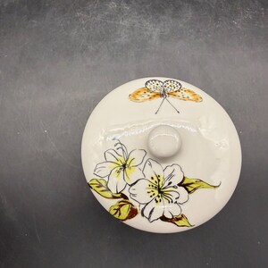 May include: A round, off-white ceramic lid with a small, round knob. The lid is decorated with a butterfly and floral design. The butterfly has orange and black wings. The flowers are white with yellow centers and green leaves.