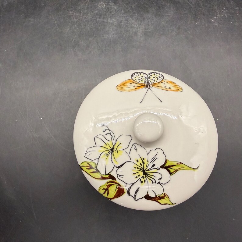May include: A round, off-white ceramic lid with a small, round knob. The lid is decorated with a butterfly and floral design. The butterfly has orange and black wings. The flowers are white with yellow centers and green leaves.