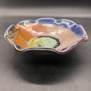 May include: A handmade ceramic bowl with a wavy rim. The bowl is glazed in shades of blue, orange, and green.