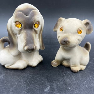 May include: Two ceramic dog figurines, one brown and white with long ears and the other gray with short ears. Both dogs have amber colored glass eyes.