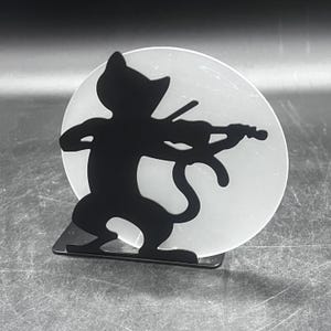 May include: A black metal cat silhouette playing a violin, set against a frosted, circular background. The cat is depicted in a playful pose, with the violin held in its paws. The design is mounted on a small, rectangular base.