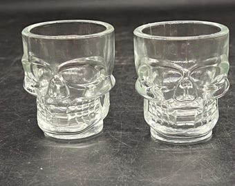 May include: Two clear glass shot glasses shaped like skulls. The glasses have a detailed skull design, including eye sockets, a nose cavity, and teeth. The shot glasses are transparent, allowing light to pass through.