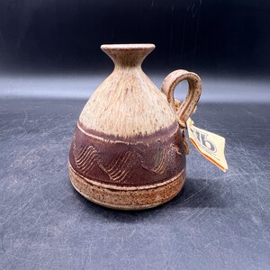 May include: A small, brown and tan ceramic vase with a handle. The vase has a wavy design around the middle and a brown glaze.