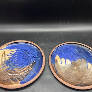 May include: Two handmade ceramic plates with a unique design. Each plate features a blue and brown glaze with a reddish-brown rim. The blue glaze has a swirl pattern, and the brown glaze has a drip effect.