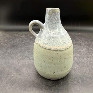 May include: A ceramic jug featuring a two-tone glaze with a light sage green base speckled with brown dots and a textured, mottled light blue upper section. The vessel has a rounded body, a narrow neck, and a single curved side handle.