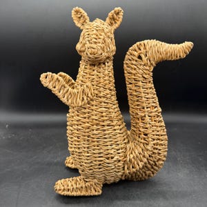 May include: A woven squirrel figurine in shades of tan and brown. The squirrel is standing upright with its arms outstretched and its tail curved. The figurine is made of woven material, giving it a textured appearance.