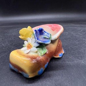 Peut inclure: Un pot de fleurs en céramique en forme de chaussure, jaune, marron et rose. Il est orné de fleurs jaunes, bleues et blanches. La chaussure a des accents bleus sur le talon.