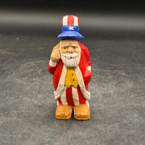 May include: A carved wooden figurine of Uncle Sam, wearing a red and white striped coat, matching trousers, and a blue hat with white stars. The figure has a white beard and is holding a sack. The figurine is standing on a dark surface.