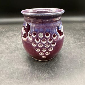 May include: A purple ceramic candle holder with a grape cluster design. The holder has a round shape with cut-out circles and leaf shapes. The top rim is a darker shade of purple.