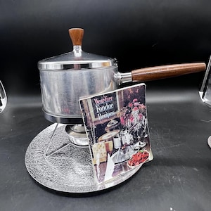 May include: A vintage fondue pot set with a silver-colored pot, wooden handle, and a matching stand. The set includes a recipe booklet titled "Wear-Ever Fondue Recipes". The pot has a wooden knob on the lid.