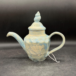 May include: Ceramic teapot with a seashell design. The teapot is primarily cream-colored with blue accents. The body features a large seashell design, and the lid is topped with a spiral seashell. The spout and handle are also blue.