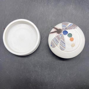 May include: White ceramic trinket box featuring a dragonfly design. The dragonfly is crafted from colourful ceramic pieces and has a brown body, blue wings, and green and orange accents.