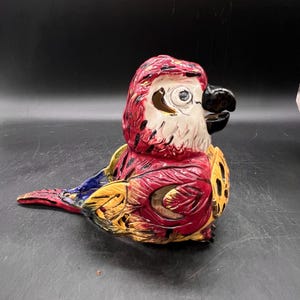 May include: A ceramic parrot figurine with red, yellow, blue, and white paint. The parrot is sitting on its tail feathers and has its head turned to the side.