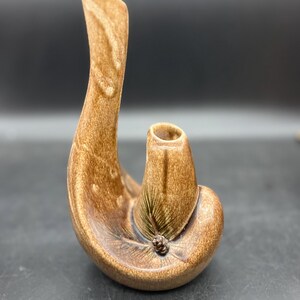 May include: A brown ceramic vase with a unique, curved design. The vase has a tall, slender neck that curves gracefully. The body of the vase has a textured surface and a small, decorative element near the base. The vase is a warm, earthy brown color.